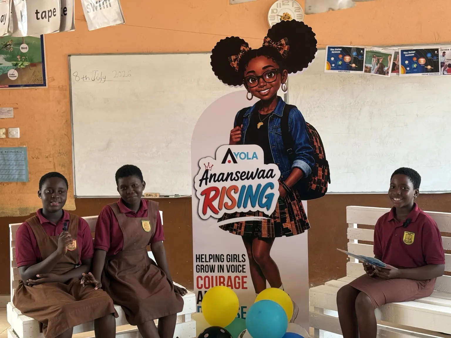 Three girls seated beside Ayola's girls' confidence programme display in their classroom.